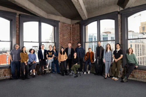 15 members of the CAST Team pose and smile for a photo in their office at Warfield Commons. A tall man stands in the corner of the room while other employees are staggered, sitting on chairs or standing on both sides of the man in the corner.