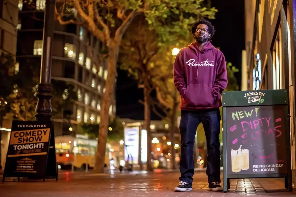 Stroy Moyd stands with his hands in the pockets of his hoodie next to an A-Frame sign for his comedy club. He looks forward and down at the camera. Market Street is blurred in the background.