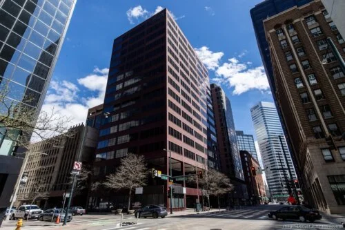 An image taken diagonally across from a roughly 15-story modern brown office building: 475 17th Street in Denver, CO. Other office buildings are seen on either side of the main building. A blue sky with intermittent clouds is above.