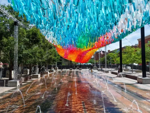 Colorful strips of fabric hang from a large aerial art installation taking up the top half of the photo. The bottom half of the photo shows the splash pond in the center of the city square that the installation hangs over.